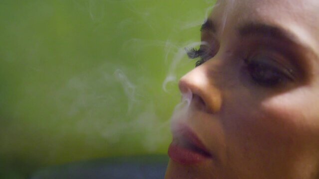 Woman against a green wall blows out smoke from a vaporizer and looks at the camera 