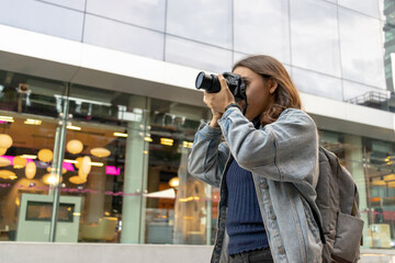 American shot of young female photographer with jean jacket and photographer backpack. He covers his face with his professional camera while looking through the camera's viewfinder.