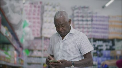 One black senior Brazilian man checking phone standing inside grocery store aisle browsing notes on smartphone device - Powered by Adobe