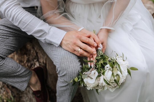 cropped photo of the bride and groom sitting on a tree stump in the forest in elegant attire. Bouquet of flowers from peonies, red shoes of the groom