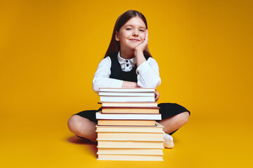 Young girl wearing uniform holding hands on stack of books while looking at camera, sitting isolated over yellow background
