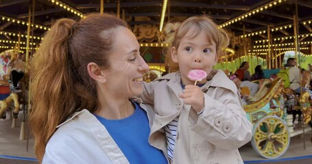 Positive little girl licks a lollipop, enjoys a sugar treat and looks at the camera. Beautiful cute baby girl eating lollipop while sitting in her mother's arms in amusement park