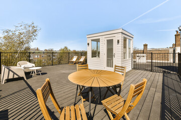 a deck with chairs and a table on the top floor, in front of a small white house there is a blue sky above