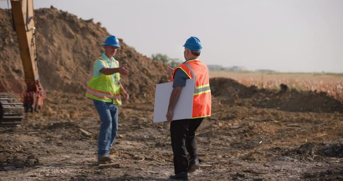 Two workers discuss building plans on a dirt jobsite with a backhoe in the background