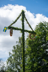 A pole and flag against green trees and blue sky. A maypole decorated, covered in flowers and leaves.