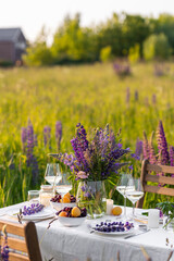 Sunset, golden hour, summer. Beautiful countryside wedding of romantic dinner decor on a blooming meadow with purple violet lupins flowers. Vase, wineglasses, cherry and apricot. Wooden table, basket