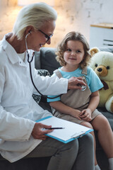 Kind senior female paediatrician doctor examining little child, cute girl during home visit or clinic check up. Concept of healthcare, medical assistance, insurance. Prescribing treatment for disease