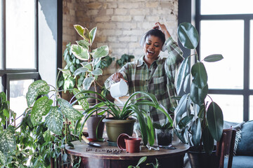 Attractive plus size African American young woman taking care about home plants. Enjoying household activity, singing, dance. Morning routine and mediation, mental health concept. Cozy green interior