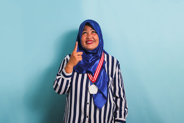 A happy middle-aged Asian woman, wearing a blue hijab and a striped shirt, is pointing her finger upwards towards the copyspace. She is wearing a medal and is isolated on a blue background