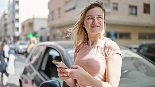 Young blonde woman using smartphone standing by car at street