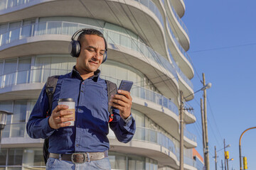 Young latin man with headphones looking at his mobile phone and holding a paper cup with coffee in...