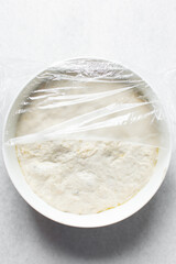 Plastic wrap covered bread dough that has doubled in size , dough that has fermented, proofed dough in a white bowl , process of making focaccia
