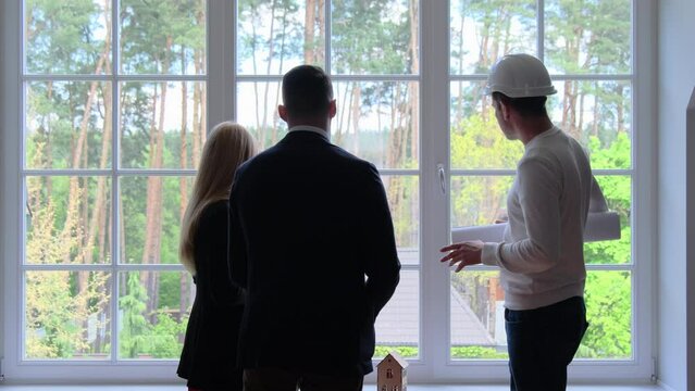 Architect in white hardhat tells rich couple about house renovation details. Builder prepares plan for summer residence renovation