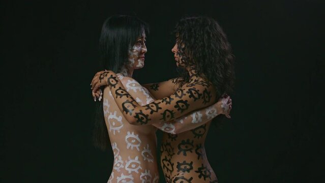 Two women with painted bodies embrace on a black backdrop