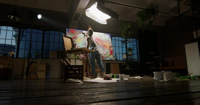 Millennial girl artist paints picture in loft studio using spatula to apply paint to canvas. Abstraction cinematic. Creative studio large picture stands on easel illuminated. Painter concept