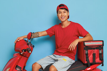 Chinese guy in red cap and red t-shirt sitting on red scooter with red delivery bag on blue...