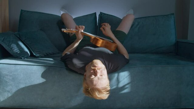 A man resting lying down. Media. A handsome man lying on a bed and playing the ukulele under the rays of the sun in a bright room.