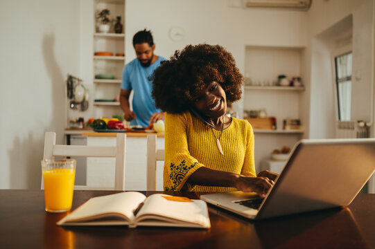 A cheerful african american female freelancer is working from home while her husband is cooking lunch.