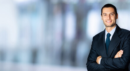 Portrait of happy confident businessman in black suit and blue tie, with crossed arms pose, copy space for some text, advertising or slogan, standing against blured office background