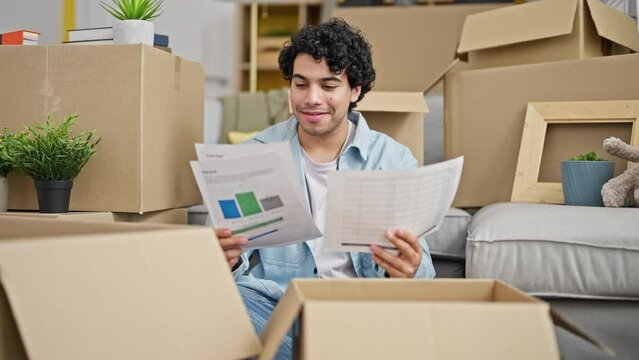 Young latin man reading document sitting on floor smiling at new home