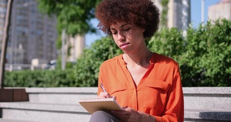 Cute attractive woman artist is sitting on stone street staircase outside writing notes in notebook or drawing something - Powered by Adobe