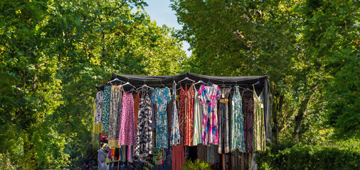 Alternative and ecological market stall with women's clothing of colorful and flowery dresses hanging from the ceiling. Surroundings of many green trees in a park and good sunlight.
