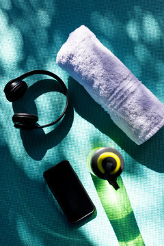 Vertical image of close up of bottle of water, towel, smartphone and headphones on mat on sunny day