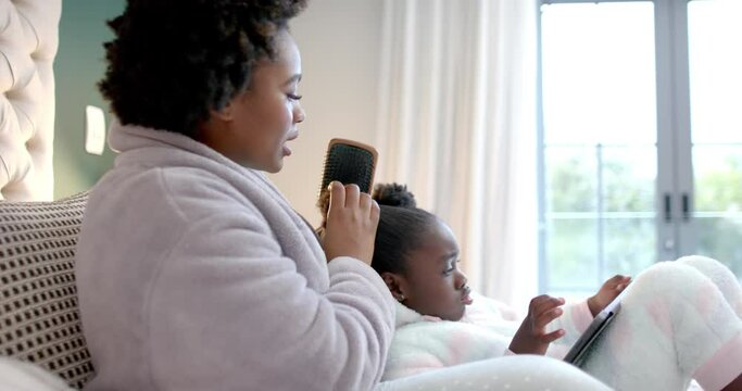 Happy african american mother combing hair of daughter using tablet in bedroom, slow motion