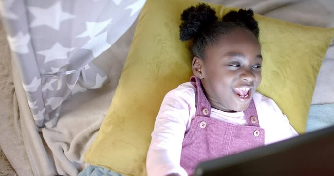 Happy african american girl lying in tent and using tablet at home, slow motion