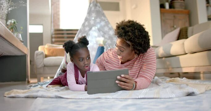 Happy african american mother and daughter lying outside tent and using tablet at home, slow motion