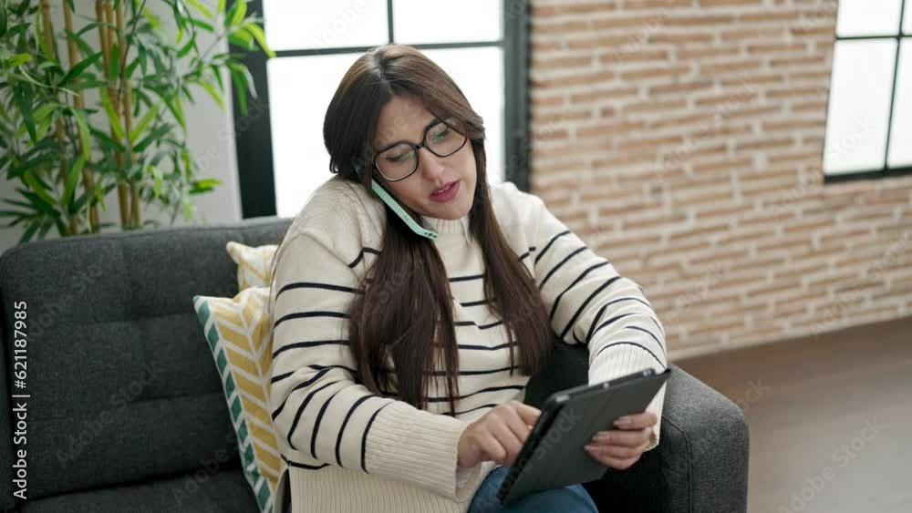 Wall mural Young beautiful hispanic woman using touchpad talking on smartphone at home