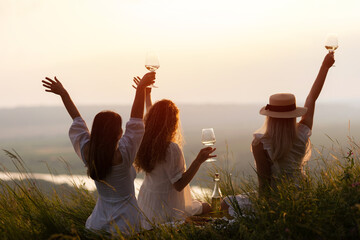 Beautiful girlfriends having picnic on the hill on summer sunset. They sitting with hands up and...