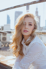 Charming young woman in white clothes with embroidery sitting near fence on sunny roof of high-rise city building.