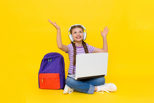 The child is engaged in an online lesson with a laptop. A young girl sits cross-legged on the floor with a backpack, headphones and looks up at your advertisement. Yellow isolated background.