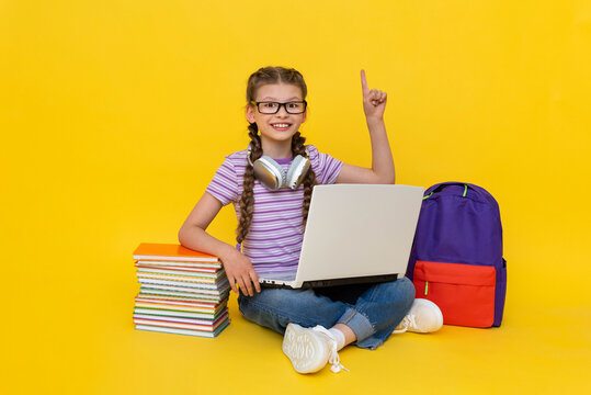 A little girl with a laptop points her finger up. Online remote education and tutoring for schoolchildren. Gadgets for school lessons. yellow isolated background.