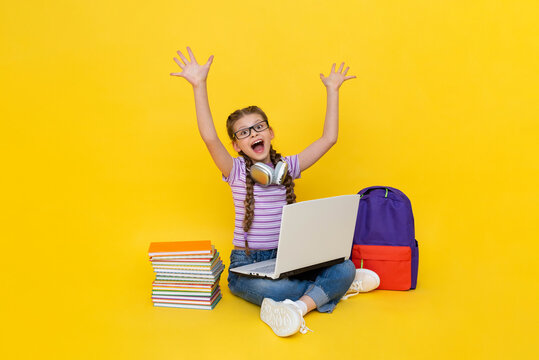 A Young Girl With A Laptop Raised Her Hands Up And Smiles Broadly, Enthusiastically. Distance Online School Education For Children. A Child With Books And A Backpack. Yellow Isolated Background.