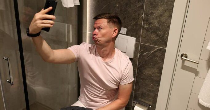 Man grimaces jokes and takes selfie toilet. Fool joking in toilet with smartphone