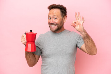 Middle age caucasian man holding coffee pot isolated on pink background showing ok sign with fingers