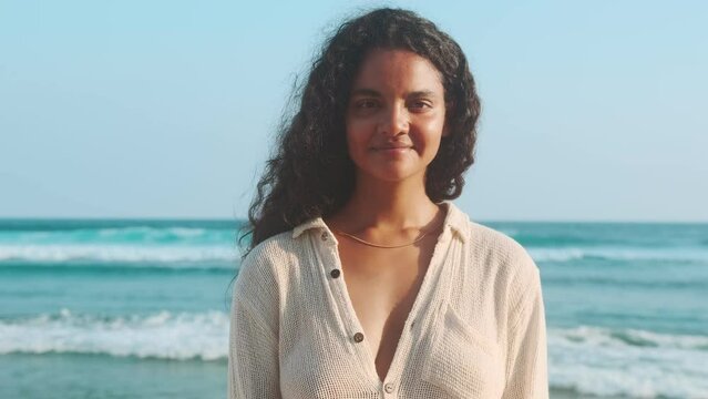 Young positive pretty Indian woman zoomer dressed in thin shirt for tropical climate smiles broadly and looks at screen recommending to use services of travel agency for trip to seaside resort.