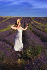 Fototapeta na wymiar A beautiful girl in a summer dress and hat stands against the backdrop of a lavender field. Pastoral. Vertical full length portrait