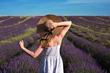 A young woman in a hat stands in the background of a lavender field. Portrait from the back of the model. Daylight