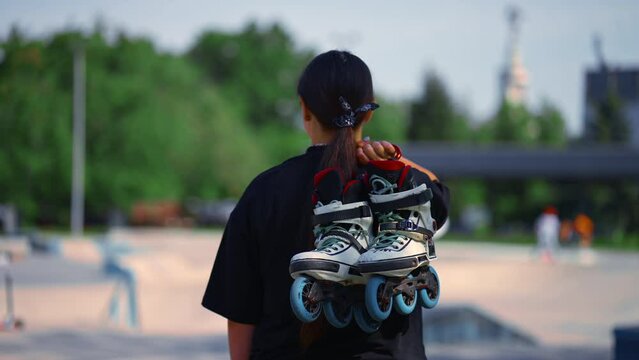 young hipster girl carries roller skates before starting training lesson on sports field street sport back view