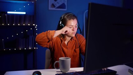 Young female gamer wear headset sitting in a dark room with cup of coffee at night and playing video games on personal computer. Gamer drinks coffee and streams game.