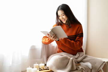 Beautiful young Asian woman reading book wearing knitted warm sweater sit on windowsill in room...