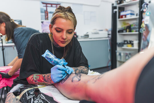 Focused young adult female tattooist drawing a tattoo on her client's leg. Blurred interior of a tattooing studio. High quality photo