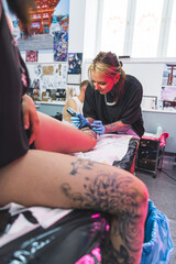 Tattooing studio concept. Interior of a tattooing studio. Vertical shot of a girl tattooist drawing a tattoo on client's leg. Blurred tattooed leg in the foreground. High quality photo