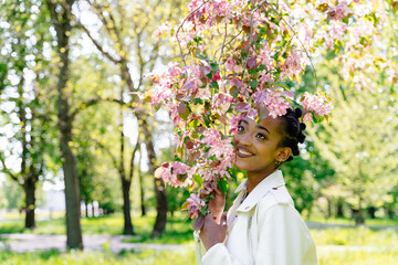 African american teenager girl with flowering branch of sakura tree in spring blooming garden enjoying warm sunny weather, serene people and mental wellbeing concept.