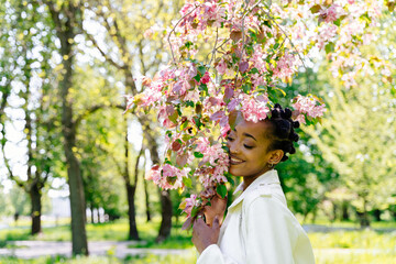 Obraz premium Concept of relaxation and mental health. African american teenager girl with flowering branch of sakura tree in spring blooming garden enjoying warm sunny weather.