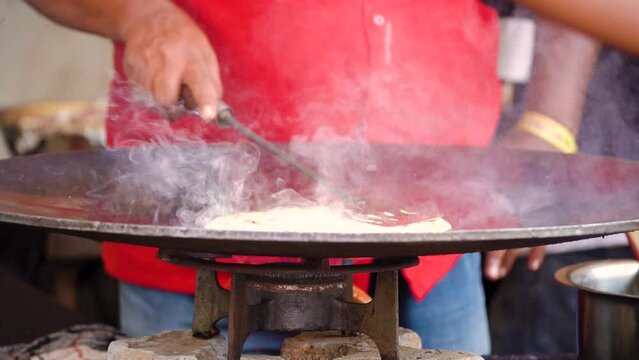 slow motion shot of man using steel spatula to make paratha flat bread on huge tawa iron pan with smoke coming from the oil and open flame on gas burner showing street food india