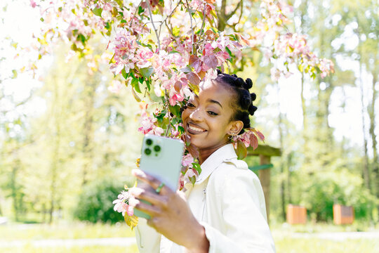 Happy smiling african young attractive woman take selfie photo with sakura branch under sakura tree in the park.
